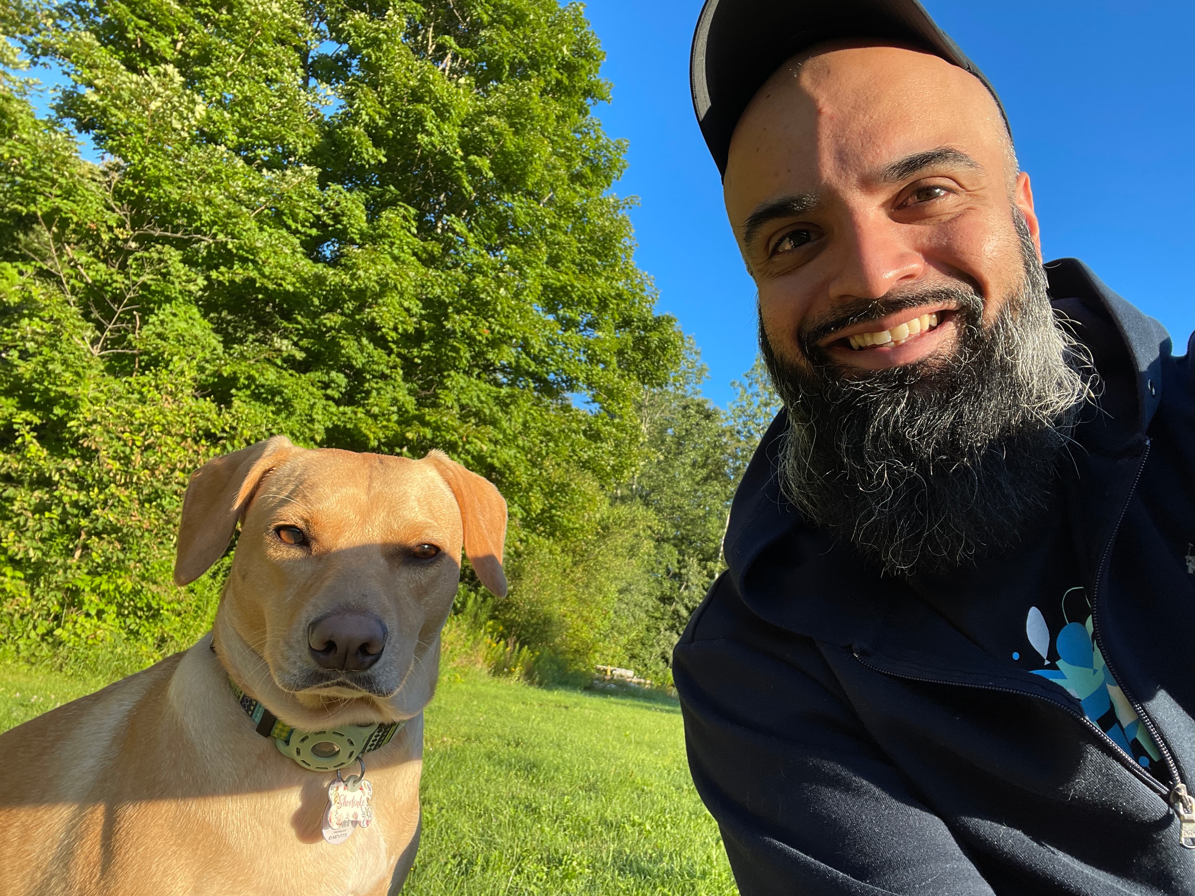 Smiling bearded man takes a selfie with a tan dog in a sunny green park.